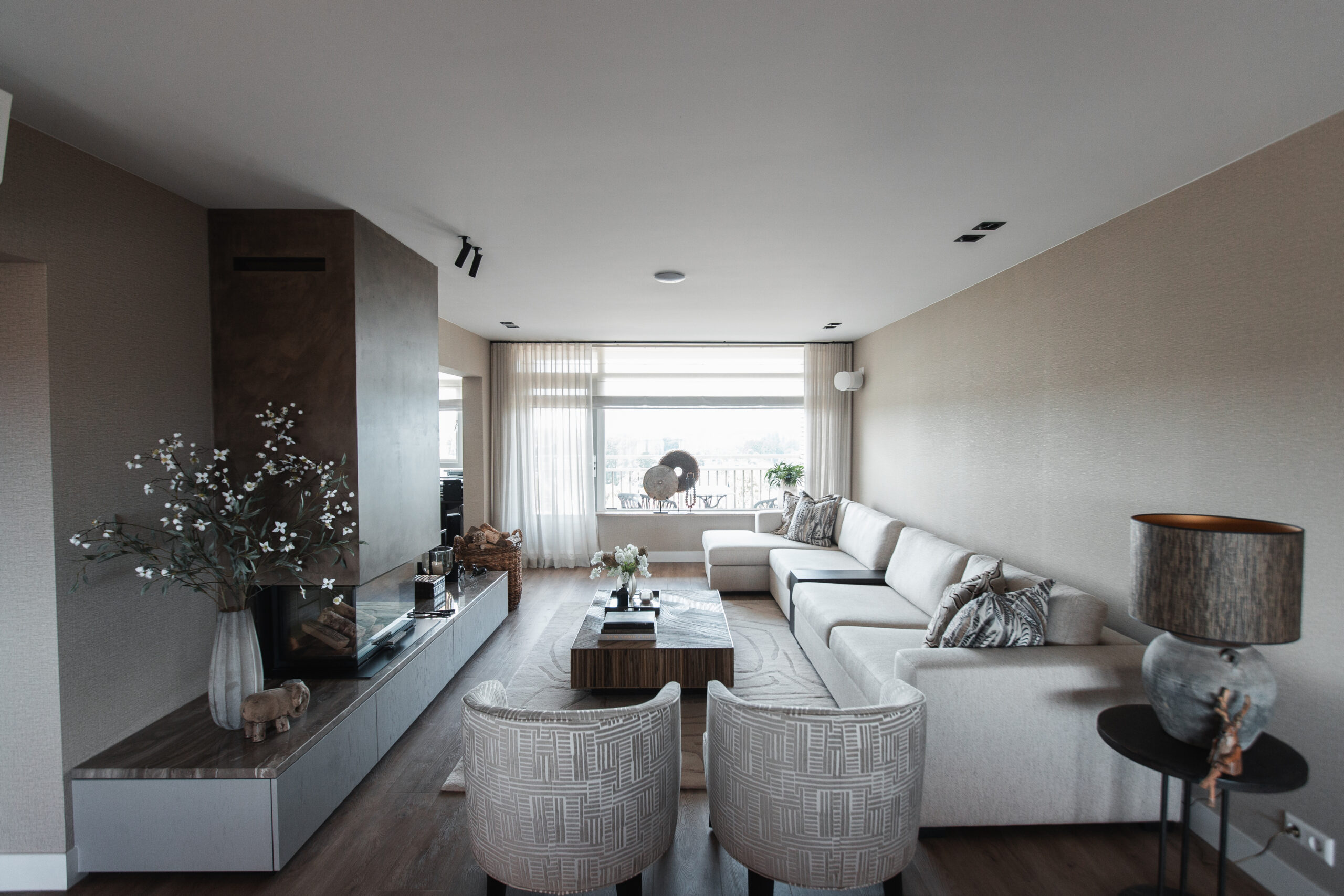 Sfeervolle woonkamer met een grote beige hoekbank, twee fauteuils, een moderne ingebouwde haard en een grote raampartij met veel lichtinval, verbouwd door DS Bouw.